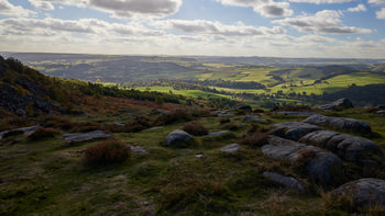 This landscape photograph captures Curbar Edge, a prominent natural feature situated in the Peak District, Derbyshire, United Kingdom. Taken during the early afternoon in the autumn season, the image showcases the striking contrast between rocky outcrops in the foreground and the patchwork of rural fields illuminated by soft, angled sunlight in the distance. The scene highlights the diverse nature of the area, with rolling hills and scattered woodland stretching across the Peak District, reflecting the scenic beauty of rural Derbyshire.